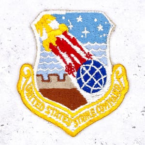 May include: Embroidered patch with a shield design and a yellow border. It shows a red and white comet heading towards a blue globe, set against a blue sky with stars and a wavy sea. The text "UNITED STATES STRIKE COMMAND" is in yellow.