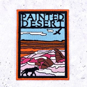 May include: A rectangular embroidered patch featuring a landscape scene with the text PAINTED DESERT at the top. The design includes a light blue sky, a dark bird silhouette, layered brown and orange hills, and pink rock formations with a black wolf.