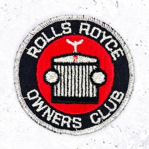 May include: Circular embroidered patch with the Rolls Royce Owners Club logo. The patch has a black border with white text: "ROLLS ROYCE OWNERS CLUB." The center features a red circle with a white and black car emblem.