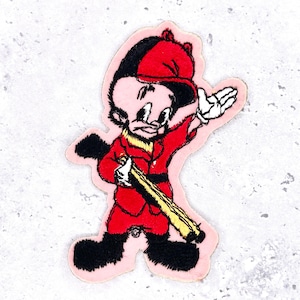 Op de afbeelding: Geborduurde patch met een stripfiguur in een rode outfit en hoed, zwarte schoenen en een geel en bruin geweer. Het personage staat tegen een roze achtergrond met een witte hand omhoog. De patch heeft een vintage esthetiek.