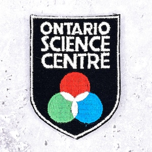 Pode incluir: Um patch bordado preto com o texto "ONTARIO SCIENCE CENTRE" em branco. Abaixo do texto, três círculos sobrepostos em vermelho, verde e azul. O patch tem uma borda branca.