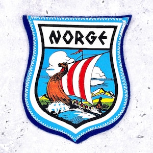 May include: A shield-shaped embroidered patch featuring a Viking ship with a red and white striped sail on blue water. The word NORGE is printed in black letters on a white background at the top. The patch has a light blue border with white stitching.