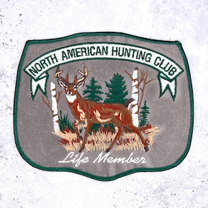 May include: Embroidered patch featuring a deer in a forest scene. The patch has the text "NORTH AMERICAN HUNTING CLUB" and "Life Member". The patch is gray with green and brown accents.