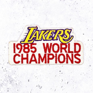 May include: Embroidered patch featuring the text "LAKERS" in yellow and purple, above "1985 WORLD CHAMPIONS" in red. The patch has a white background and a rectangular shape, perfect for apparel or accessories.