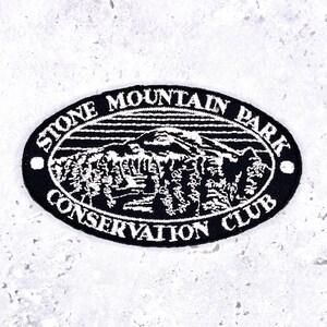 May include: An oval-shaped embroidered patch featuring a black background with white stitching. The design depicts a mountain landscape with trees and the text STONE MOUNTAIN PARK CONSERVATION CLUB embroidered in white around the border.