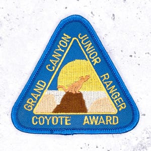 May include: Embroidered blue triangular patch with the text "GRAND CANYON JUNIOR RANGER COYOTE AWARD" in gold. The patch features a coyote on a brown rock formation with a yellow sun in the background.
