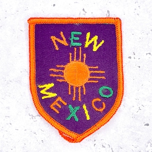 Op de afbeelding: Geborduurde patch met de staat New Mexico. De patch heeft een paarse achtergrond met de woorden "NEW MEXICO" in geel, groen en oranje. Een oranje zonnesymbool staat in het midden en de patch is omzoomd met oranje stiksels.