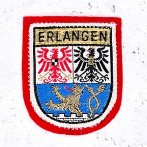 May include: Embroidered patch with the word "ERLANGEN" in gold lettering. The patch features a shield design with a red border, displaying a coat of arms with eagles and a lion in blue.