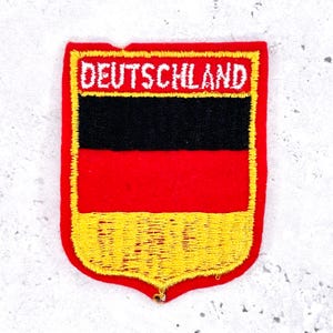 Peut inclure: Écusson brodé représentant le drapeau allemand. Il présente un fond rouge, une bande noire au centre et une section inférieure jaune. Le mot "DEUTSCHLAND" est brodé en blanc en haut de l'écusson.
