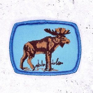 May include: Embroidered patch featuring a brown moose with large antlers, standing on green grass against a light blue background. The patch has a square shape with rounded corners and a blue border.