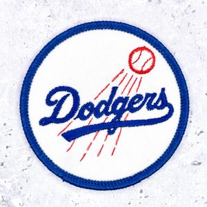 May include: A circular embroidered patch featuring a white background with a blue border. The text Dodgers is stitched in blue script, accompanied by a red baseball graphic with red motion lines trailing behind it across the white surface.