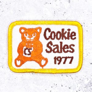 Op de afbeelding: Geborduurde patch met een witte achtergrond en een gele rand. De patch toont een bruine beer met een koekje, met de woorden "Cookie Sales 1977" in bruine tekst.
