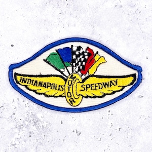 May include: An embroidered patch featuring the Indianapolis Speedway logo with yellow wings, a central yellow wheel, and a cluster of green, blue, black and white checkered, red, and yellow flags. The patch has a blue border and white background.