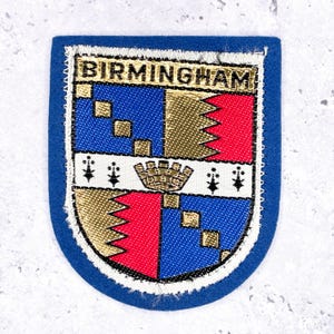May include: A blue, embroidered patch with the word "BIRMINGHAM" at the top. The patch features a shield design with blue, red, and gold sections, and a crown emblem. The patch is bordered with a blue fabric.
