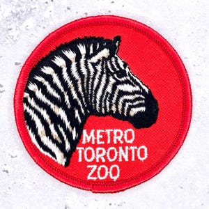 May include: A circular embroidered patch featuring a black and white zebra head profile on a solid red background. The text METRO TORONTO ZOO is stitched in white capital letters below the zebra. The patch has a red stitched border edge.