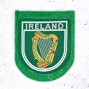 May include: A green patch featuring the word "IRELAND" above a gold harp emblem. The harp is set against a green background with white and green borders. The patch has a green felt-like border.