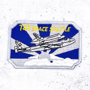 May include: Embroidered patch depicting a 747 aircraft with a space shuttle on top. The patch has a blue background with white rays and yellow text reading "747 SPACE SHUTTLE". The border is silver.