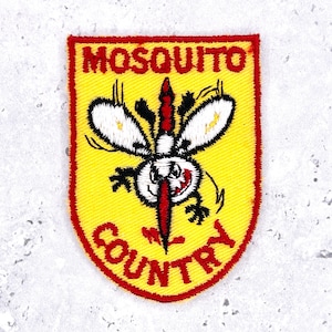 Op de afbeelding: Geborduurde patch met een gele achtergrond en een rode rand. De patch toont een cartoon mug met witte vleugels, zwarte poten en een rode steeksnuit. De woorden "MOSQUITO COUNTRY" zijn rood.