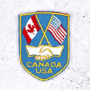 May include: A blue patch with a yellow border features the flags of Canada and the United States crossed above a handshake. The text "CANADA USA" is in yellow. The Canadian flag is red and white, and the US flag is red, white, and blue.
