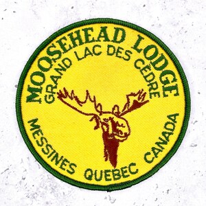 May include: A circular embroidered patch with a yellow background and a dark green border. It features a brown moose head illustration in the centre and the text MOOSEHEAD LODGE, GRAND LAC DES CEDRE, MESSINES QUEBEC CANADA in dark green lettering.