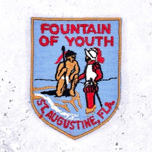 May include: Embroidered patch with a light blue background and a gold border. The patch features the text "FOUNTAIN OF YOUTH" in red, and "ST. AUGUSTINE, FLA" in red. The patch depicts two figures, one holding a spear.