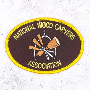 May include: An oval patch with the text "NATIONAL WOOD CARVERS ASSOCIATION" in gold lettering. The patch has a brown background with a gold border. The centre features a wood carving tool design.