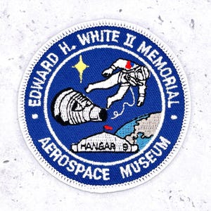 Pode incluir: Patch bordado circular com fundo azul. Apresenta um astronauta, uma cápsula espacial e um mapa da Terra. O texto diz "EDWARD H. WHITE II MEMORIAL AEROSPACE MUSEUM". Há também uma estrela amarela.