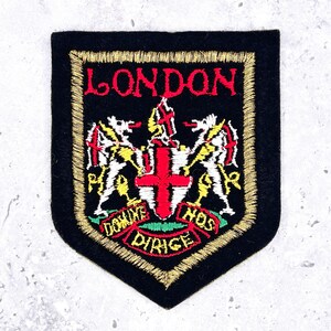 May include: A black shield-shaped embroidered patch featuring the word LONDON in red at the top. The center displays a white shield with a red cross, flanked by two white dragon-like figures, with a red banner below reading DOMINE NOS DIRIGE.