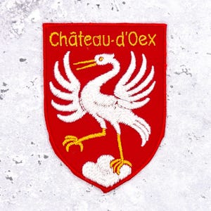 Könnte beinhalten: Gestickter Aufnäher mit rotem Schild, das einen weißen Storch und den Text "Château-d'Oex" in Gelb zeigt. Der Storch hat gelbe Beine und einen gelben Schnabel, mit ausgebreiteten Flügeln. Der Aufnäher ist wahrscheinlich ein Souvenir oder ein Emblem.
