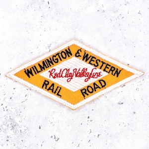 May include: A diamond-shaped embroidered patch with a yellow border and a white center. The black text reads WILMINGTON & WESTERN RAIL ROAD, while the red script text in the center reads Red Clay Valley Line. The patch has a stitched edge.