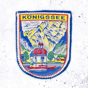 May include: A woven patch with a blue border and a white, red, and yellow design. The patch features the text "KONIGSSEE" at the top, with a mountain landscape, a church, and a boat on a lake.