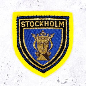 Peut inclure: Un écusson brodé en forme de bouclier avec une bordure jaune vif et un blason bleu central. Le blason présente une couronne dorée et un visage stylisé, avec le mot STOCKHOLM brodé en lettres dorées sur la partie supérieure de l'écusson.
