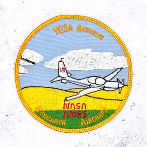 Puede incluir: Parche bordado con borde amarillo, que representa un avión blanco y azul sobre un cielo azul con nubes. El texto incluye "YO3A Acoustic" y "NASA Ames Aircraft Research". El paisaje es amarillo y verde.