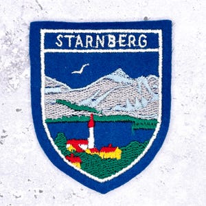 May include: A blue, shield-shaped embroidered patch with the word "STARNBERG" in white. The patch features a landscape scene with mountains, a lake, and a lighthouse. A white bird flies in the sky. The patch has a blue border.