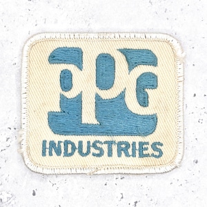 May include: A rectangular embroidered patch with a cream-colored fabric base and a stitched border. The design features a large, stylized blue monogram logo above the blue embroidered text "INDUSTRIES" in a bold, sans-serif typeface.