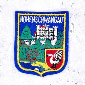 May include: Embroidered patch with a blue border and a yellow outline. The patch features the text "HOHENSCHWANGAU" at the top. The design includes a white castle, green trees, and a red and white emblem with a swan.