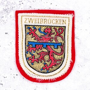 Peut inclure: Un écusson brodé en forme de bouclier avec une bordure rouge. Le centre présente un champ doré avec un lion rouge, une barre horizontale bleue et le texte ZWEIBRÜCKEN en fil doré en haut. Le motif est souligné par une couture dorée métallique.