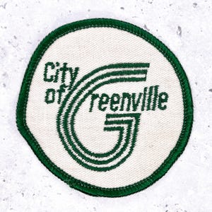 Op de afbeelding: Geborduurde ronde patch met de tekst "City of Greenville" in groen op een witte achtergrond. Het ontwerp bevat een gestileerde groene "G" en is omrand met een groene rand. De patch is rond van vorm.