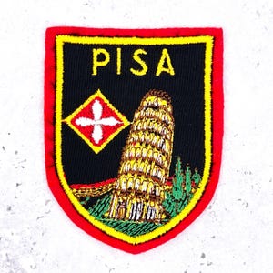 May include: A shield-shaped embroidered patch with a black background, a red border, and a yellow trim. It features the word PISA in yellow, a red and yellow diamond emblem with a white cross, and a depiction of the Leaning Tower of Pisa.