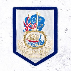 May include: A blue and white embroidered patch with a shield shape. The patch features a ship, anchor, and flags, with the text "Sault Ste. Marie Michigan FOUNDED 1668". The patch has a blue border.