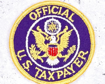 May include: A round, embroidered patch with a purple background and gold trim. The patch features the text "OFFICIAL U.S. TAXPAYER" in white, surrounding a central emblem of a golden eagle and shield.