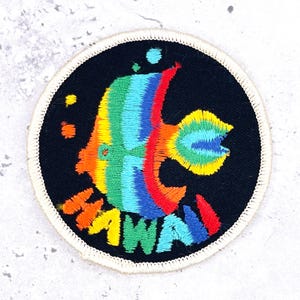 May include: A circular embroidered patch with a black background featuring a rainbow-coloured tropical fish and the word HAWAII stitched in multicoloured thread at the bottom. The patch has a white stitched border around the entire circumference.