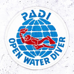May include: A circular embroidered patch featuring a white background with a blue globe graphic. A red scuba diver figure is centred over the globe. The text PADI is arched above, and OPEN WATER DIVER is arched below in blue embroidery.