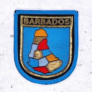 May include: A shield-shaped embroidered patch featuring the word BARBADOS in gold thread at the top. The center displays a stylized map of Barbados divided into segments of red, blue, orange, and gold thread, all bordered by a bright blue edge.