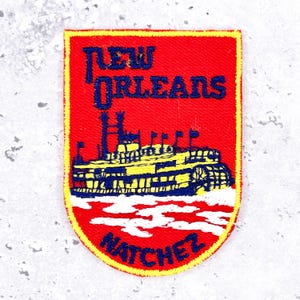 Puede incluir: Un parche bordado rectangular con borde inferior redondeado, fondo rojo y borde amarillo. Representa un barco de vapor amarillo y azul sobre olas blancas, con el texto NEW ORLEANS en la parte superior y NATCHEZ en la parte inferior.