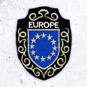 May include: Embroidered patch featuring a blue shield with a circle of gold stars, representing the European Union. The word "EUROPE" is embroidered in white above the shield. The patch has a black border with gold decorative swirls.