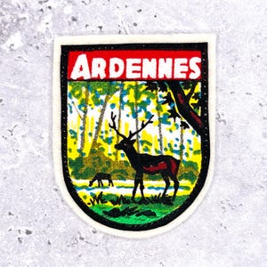 Op de afbeelding: Geborduurde patch met het woord "ARDENNES" in wit op een rode achtergrond. De patch toont een boslandschap met herten, bomen en een rivier. De patch heeft een zwarte rand en een witte rand.