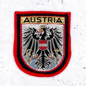 May include: Embroidered patch featuring the Austrian coat of arms. The patch has a red border, a black inner border, and a shield shape. The text "AUSTRIA" is at the top in gold lettering. The coat of arms includes a black eagle with a red and white shield.