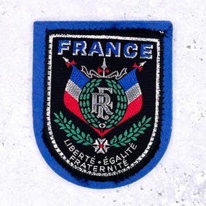 May include: A blue and black patch with the word "FRANCE" in silver lettering. The patch features the French flag colors, a wreath, and the words "LIBERTÉ, ÉGALITÉ, FRATERNITÉ".