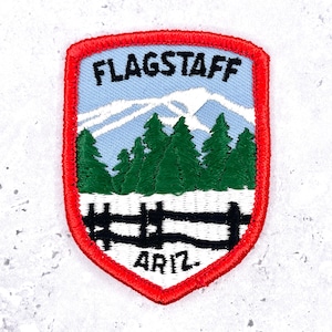 May include: A shield-shaped embroidered patch with a red border. The design features a light blue sky, white snow-capped mountains, a forest of green pine trees, and a black wooden fence over white snow. The text reads FLAGSTAFF and ARIZ.